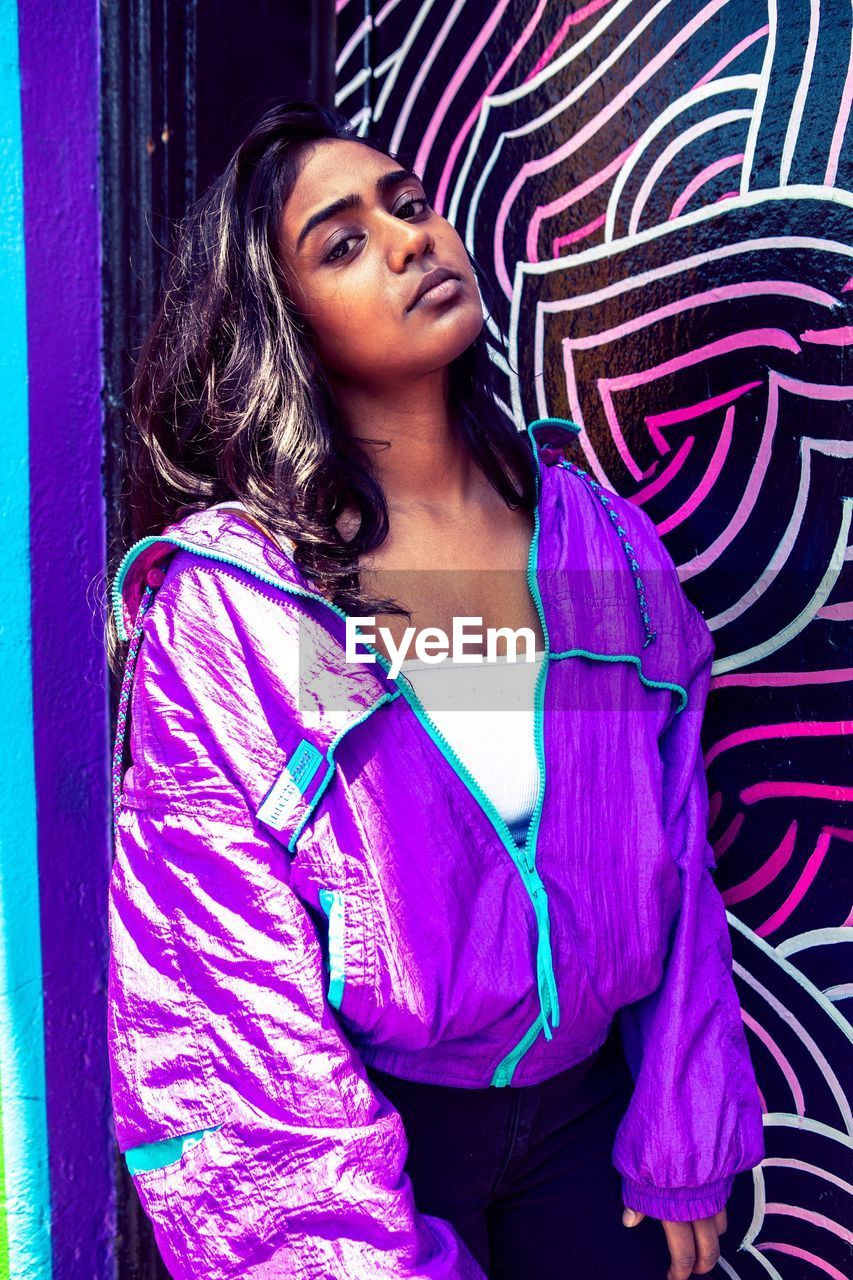 Portrait of young woman wearing purple jacket against wall