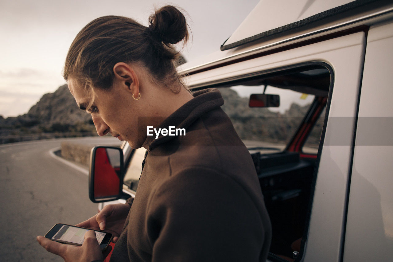 Side view of male explorer with man bun text messaging through smart phone near van on vacation