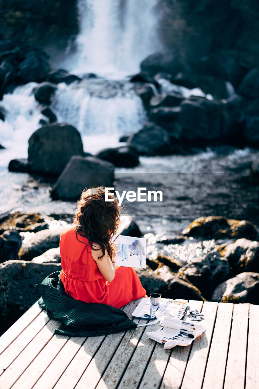 REAR VIEW OF GIRL SITTING ON ROCK AGAINST WATERFALL