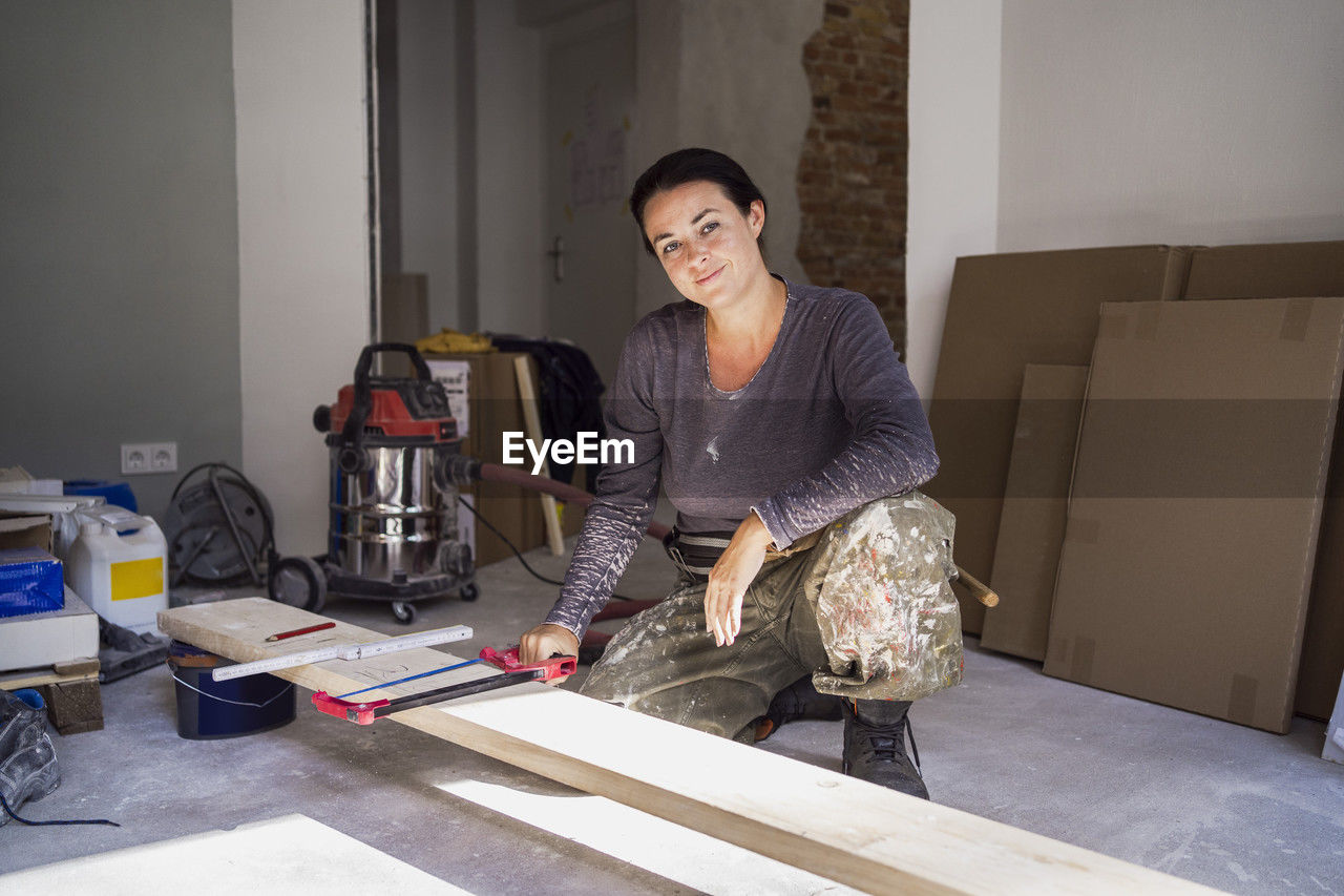 Portrait of confident female carpenter holding handsaw crouching in front of plank in apartment