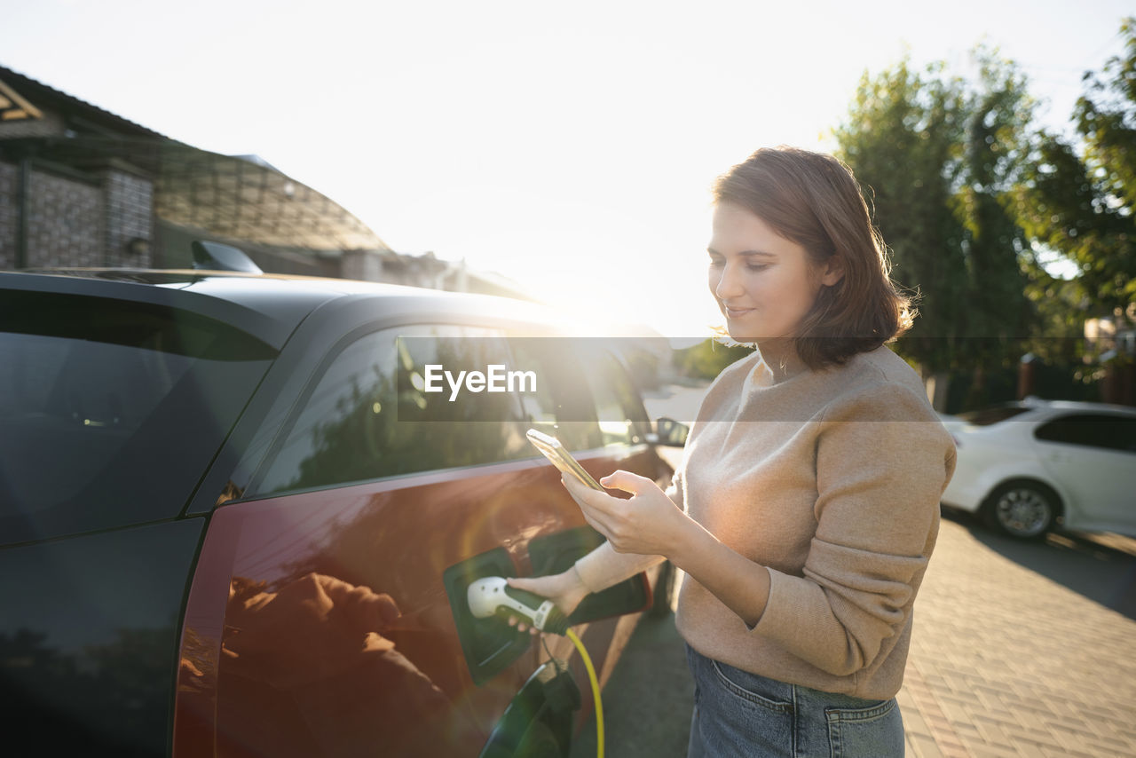 Smiling woman using smart phone and charging car on sunny day