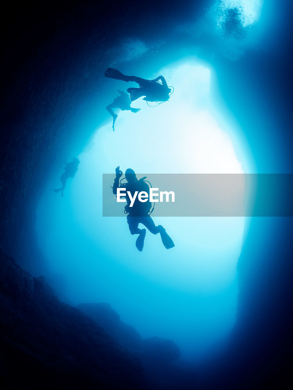 Unrecognizable professional divers with scuba and flippers swimming in narrow dark cave with rough stony formations undersea in deep clear water