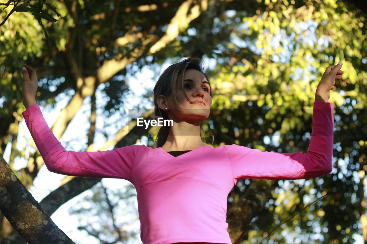 Low angle view of woman looking away with arms raised while standing against trees