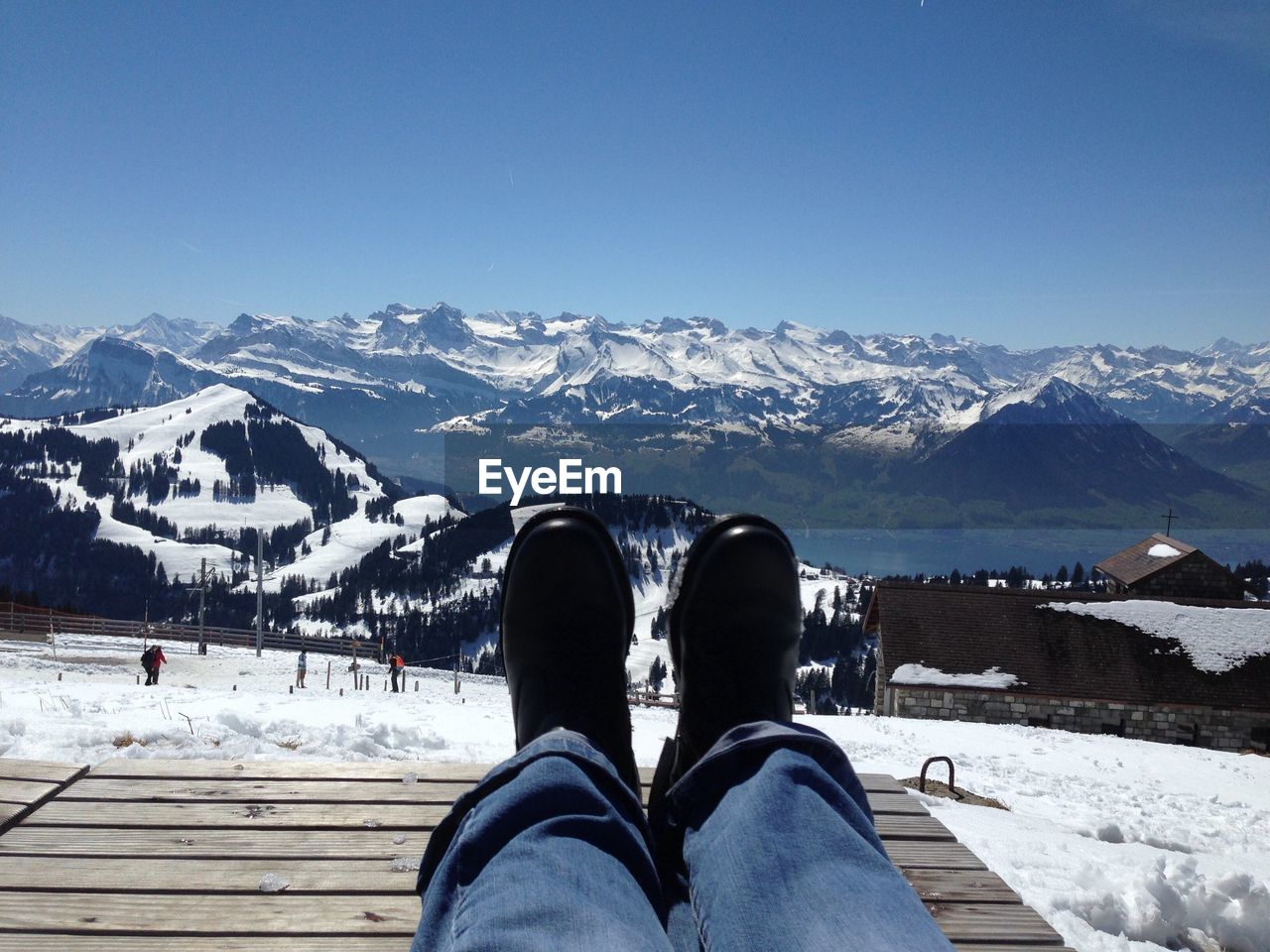 Low section of person on wooden sea at snow covered field by mountains against clear sky