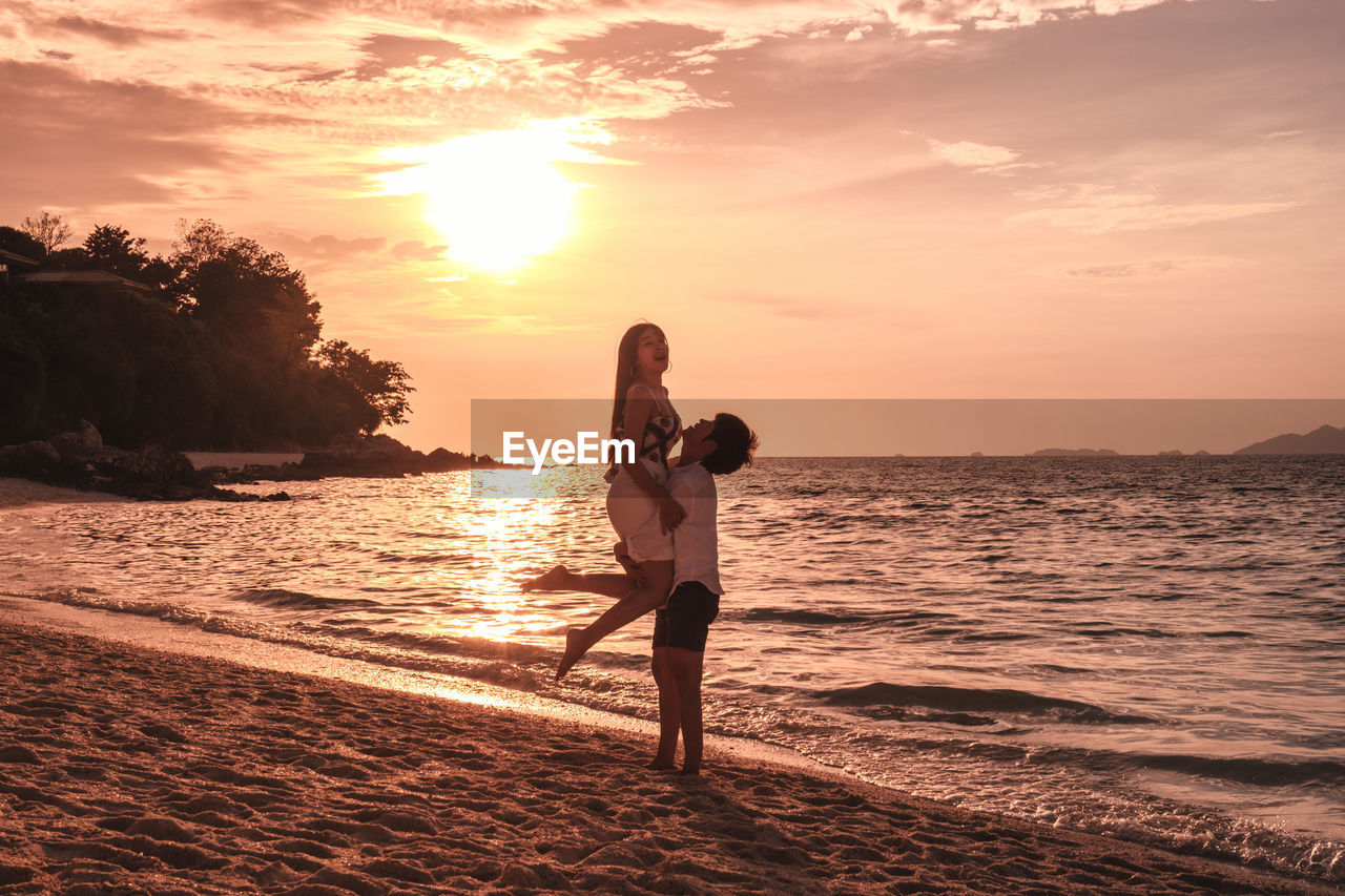 Man carrying woman while standing at beach against sky during sunset