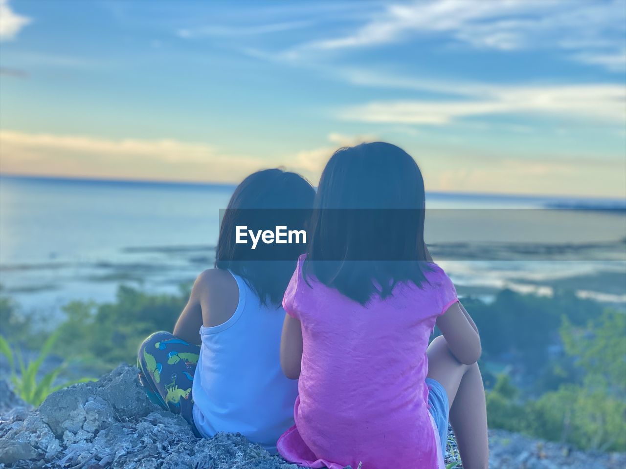 Two kids watching the sunset sitting on top of the mountain overlooking the ocean. 