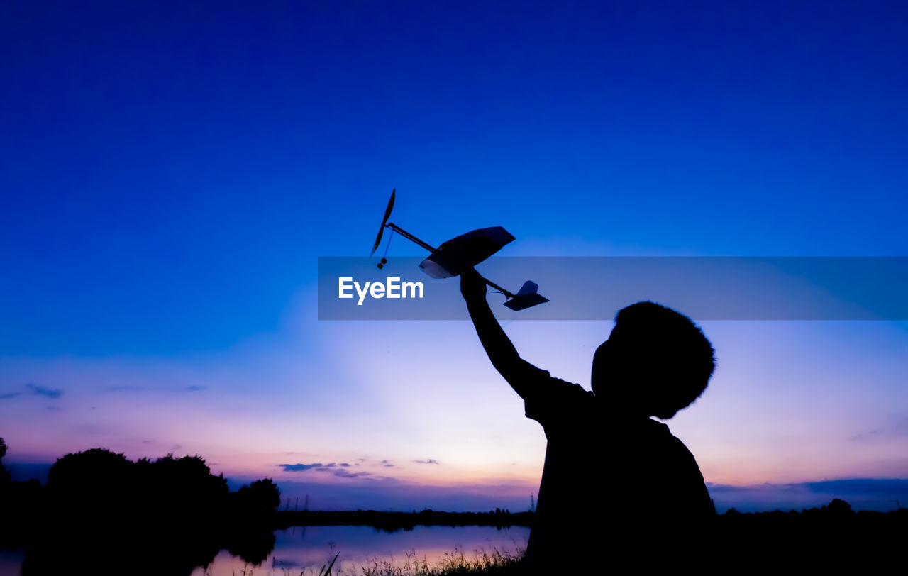 Silhouette of boy playing with toy airplane against sky