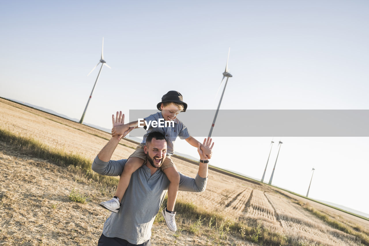 Father carrying son on shoulders in front of wind turbines