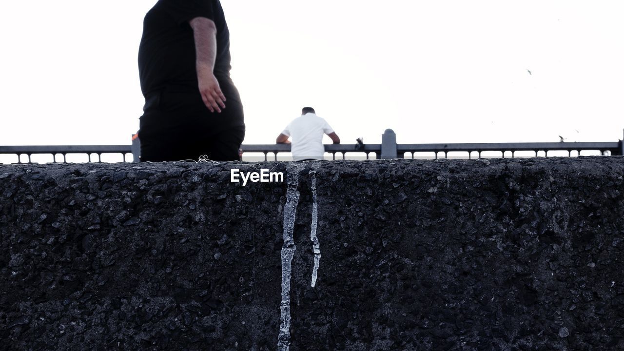 REAR VIEW OF MEN PERCHING ON RAILING AGAINST SKY