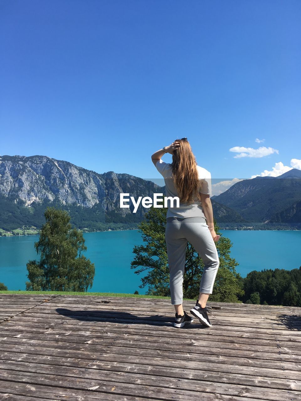 REAR VIEW OF WOMAN STANDING BY LAKE AGAINST MOUNTAIN