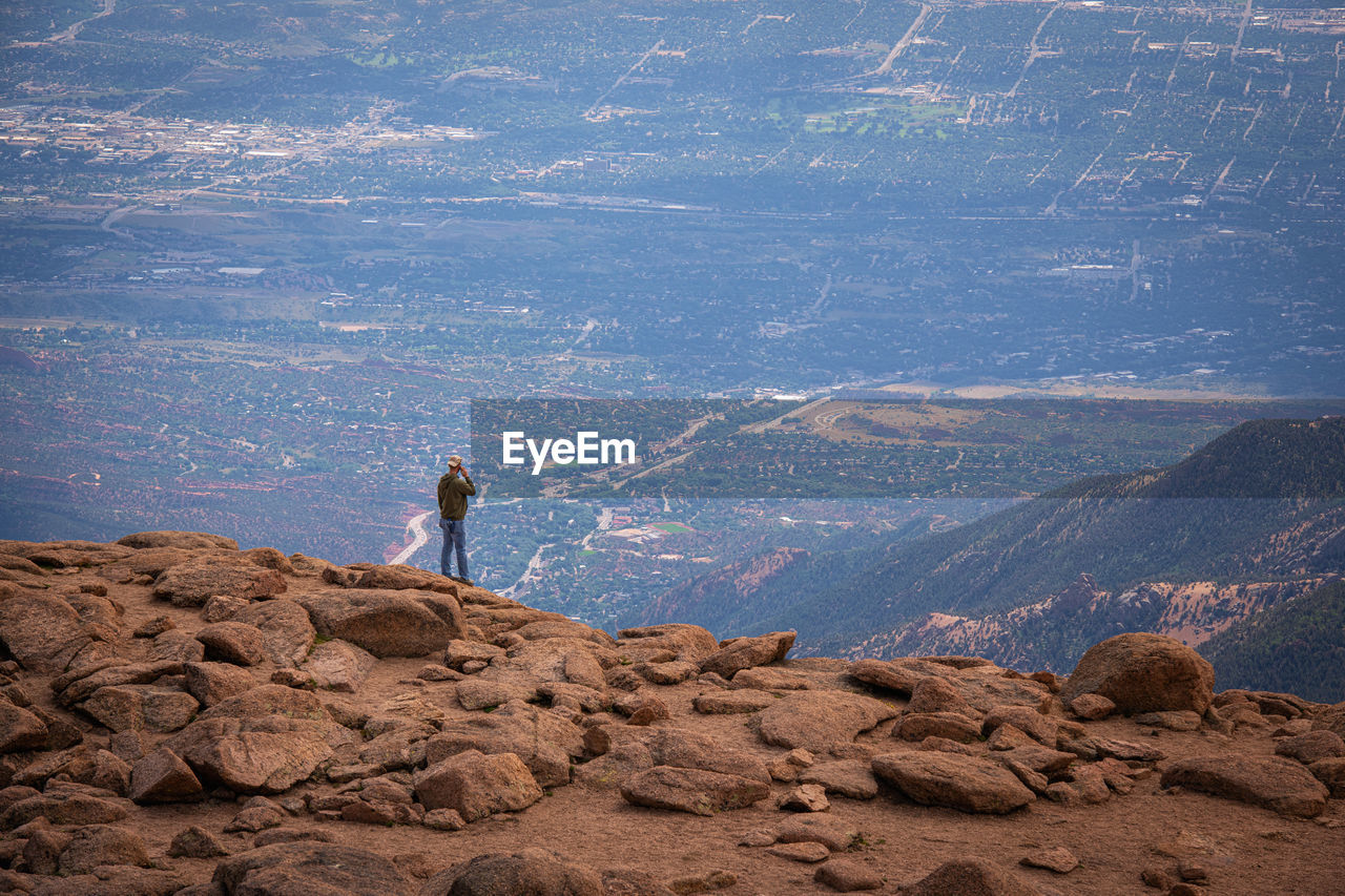 MAN STANDING ON ROCK ON LAND AGAINST MOUNTAINS