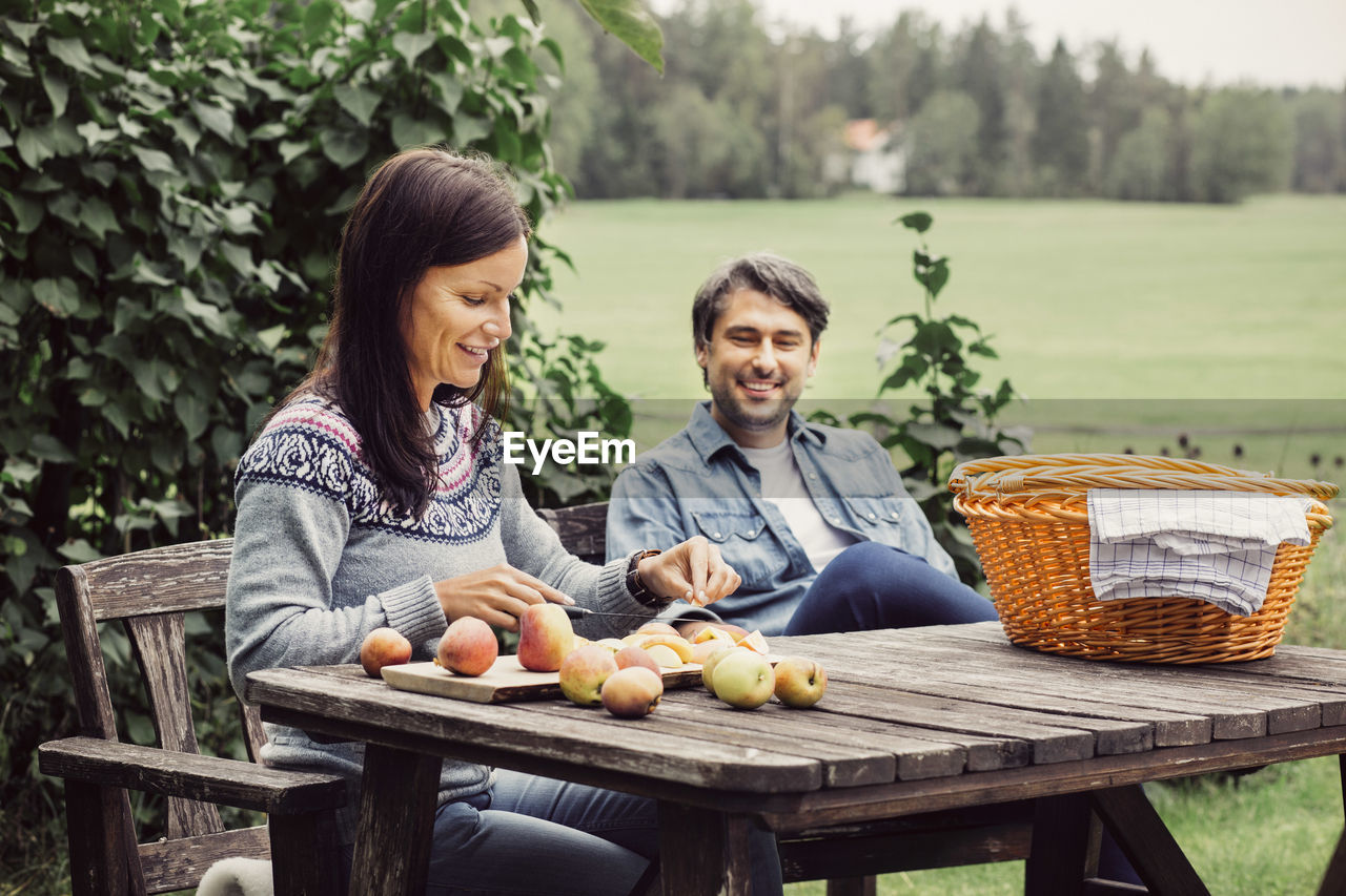 Happy man looking at woman cutting apples at table in organic farm