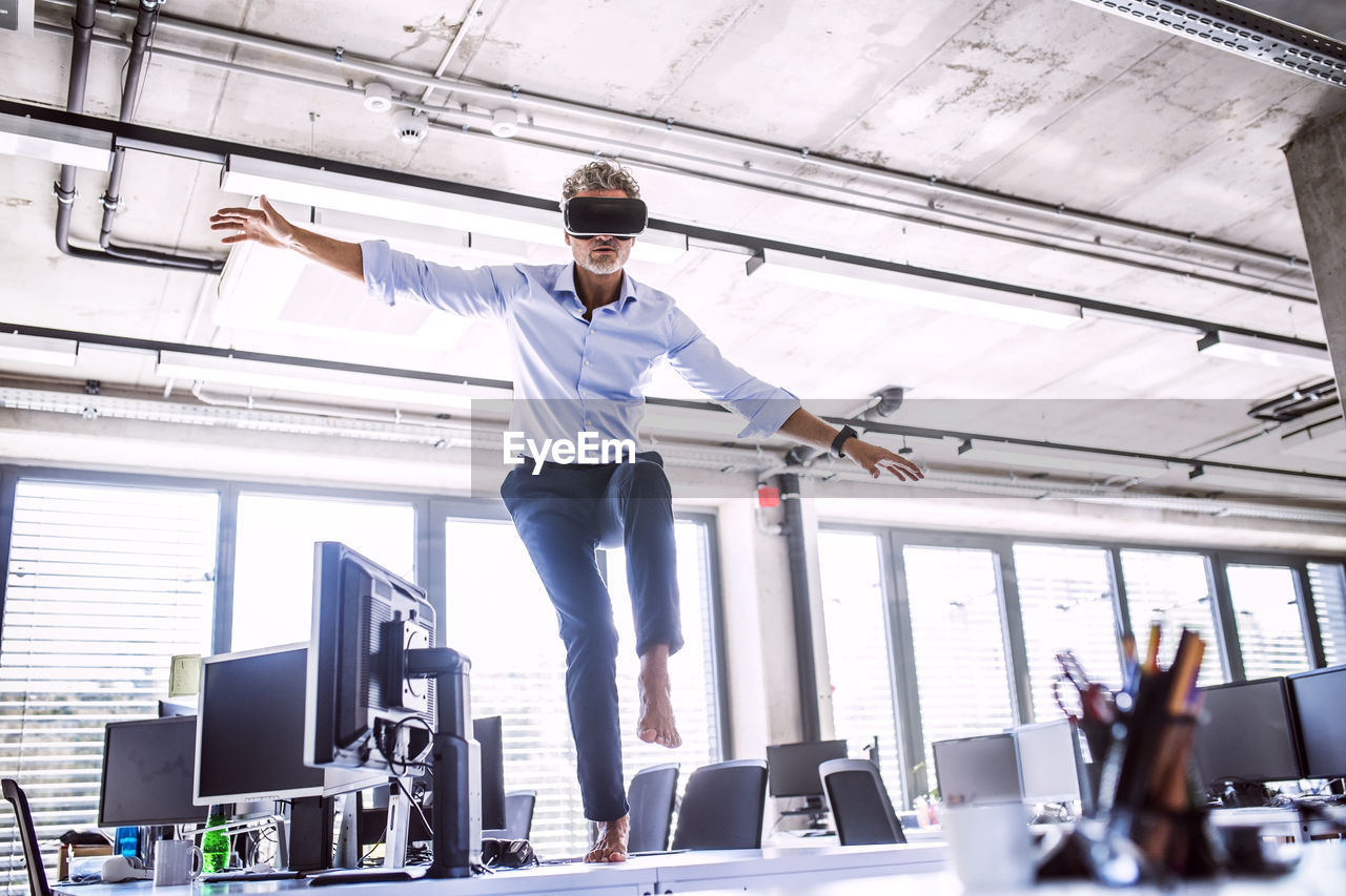 Barefoot mature businessman on desk in office wearing vr glasses barefoot