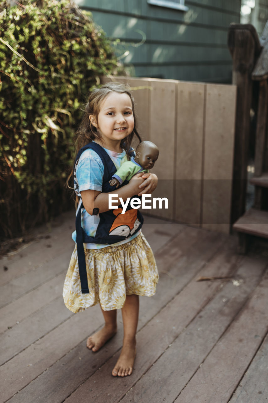 Little girl carrying babydoll in baby carrier outside in yard