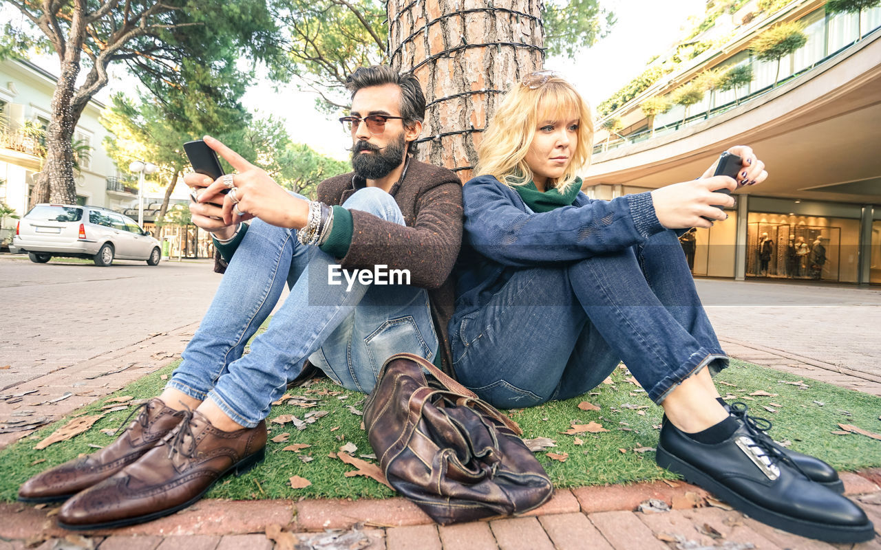 Man and woman using mobile phone while sitting against tree trunk in city