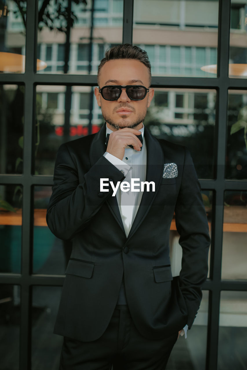 YOUNG MAN WEARING SUNGLASSES STANDING AGAINST WALL
