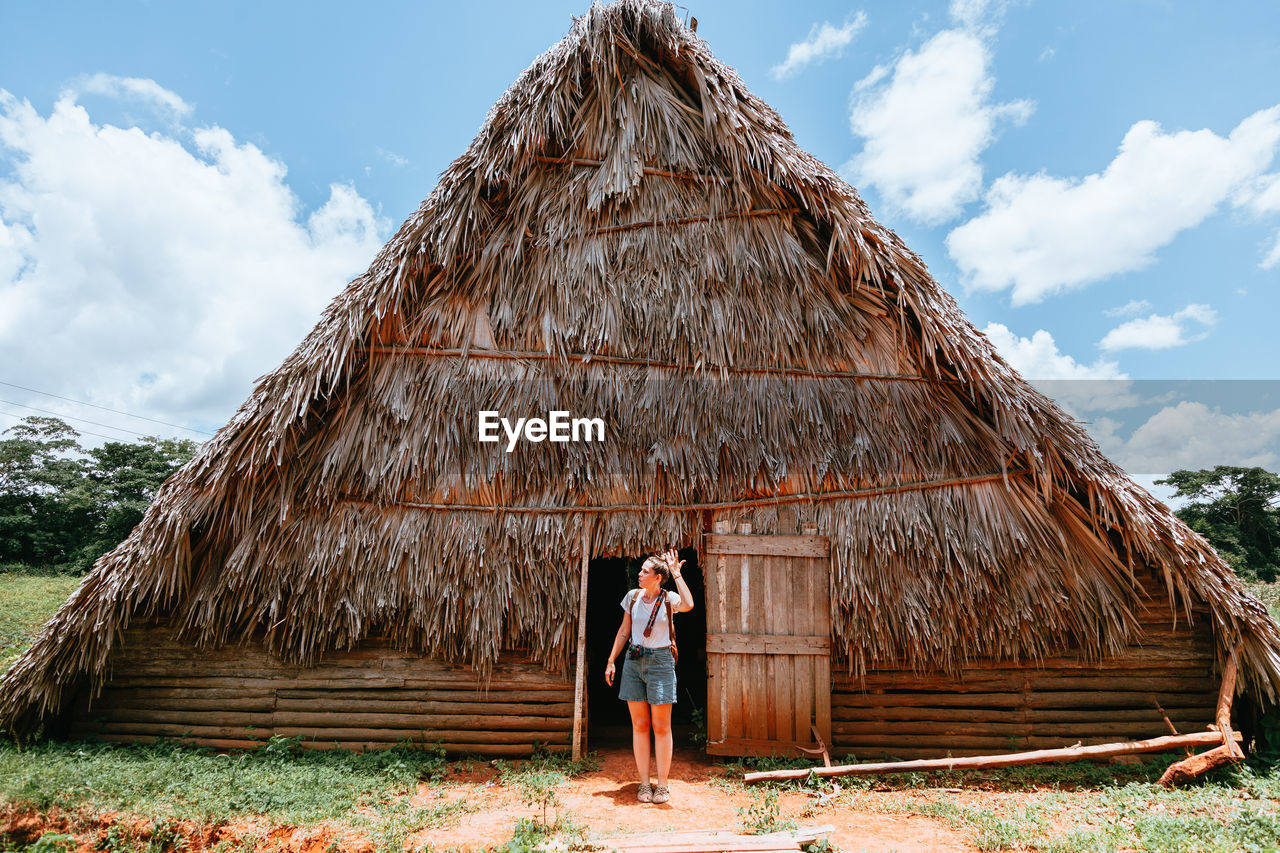 Resting lady in stylish casual clothing standing in doorway of big rural house with thatched roof and blue sky on background in cuba
