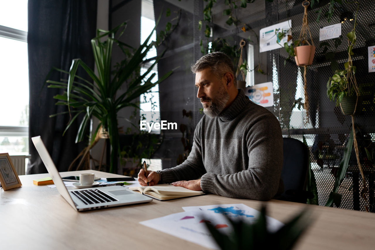 A man is sitting at a desk with a laptop and writing in a notebook