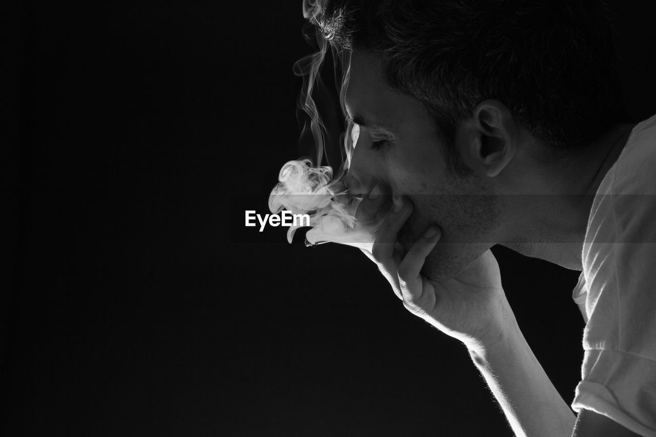 Close-up of man with hand covering mouth emitting smoke against black background