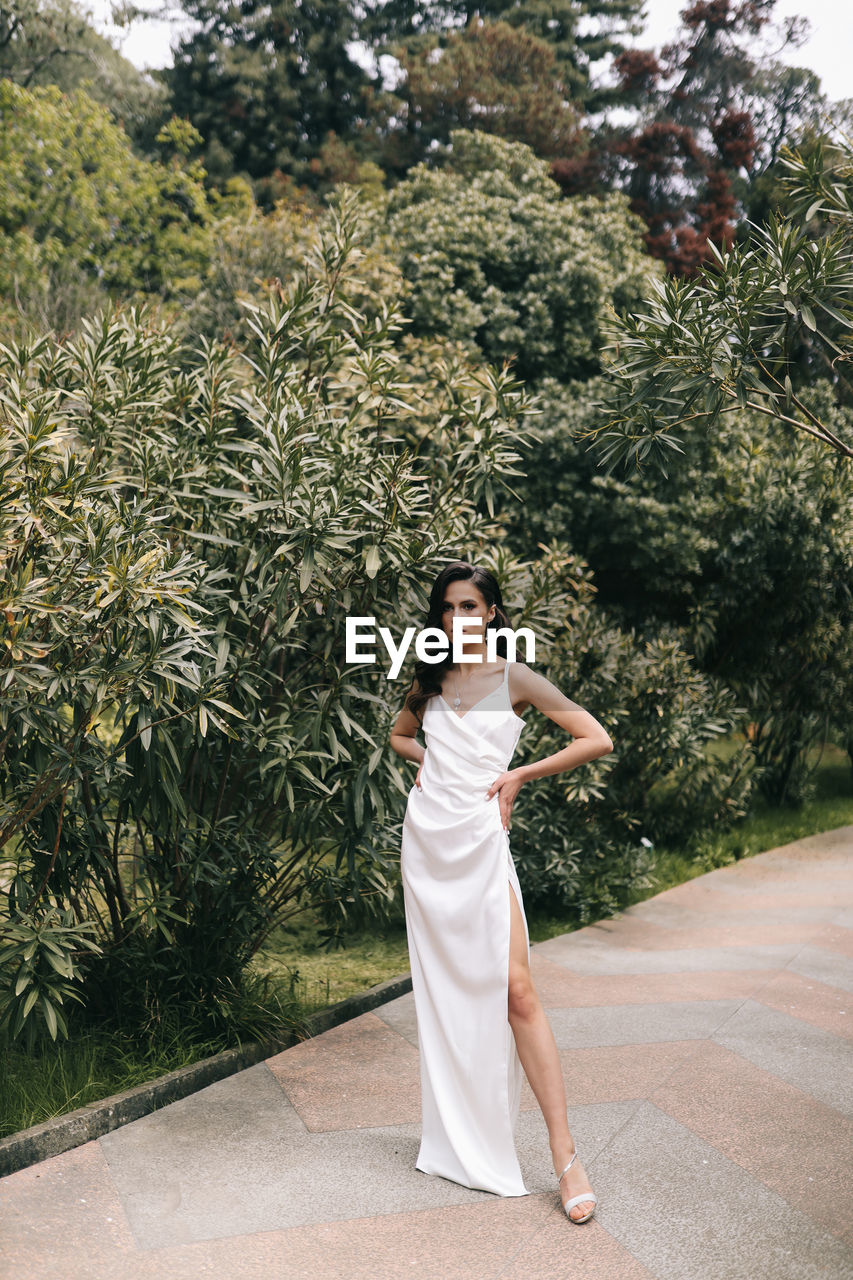 An elegant young woman bride in a wedding dress walks through a green park among plants and trees