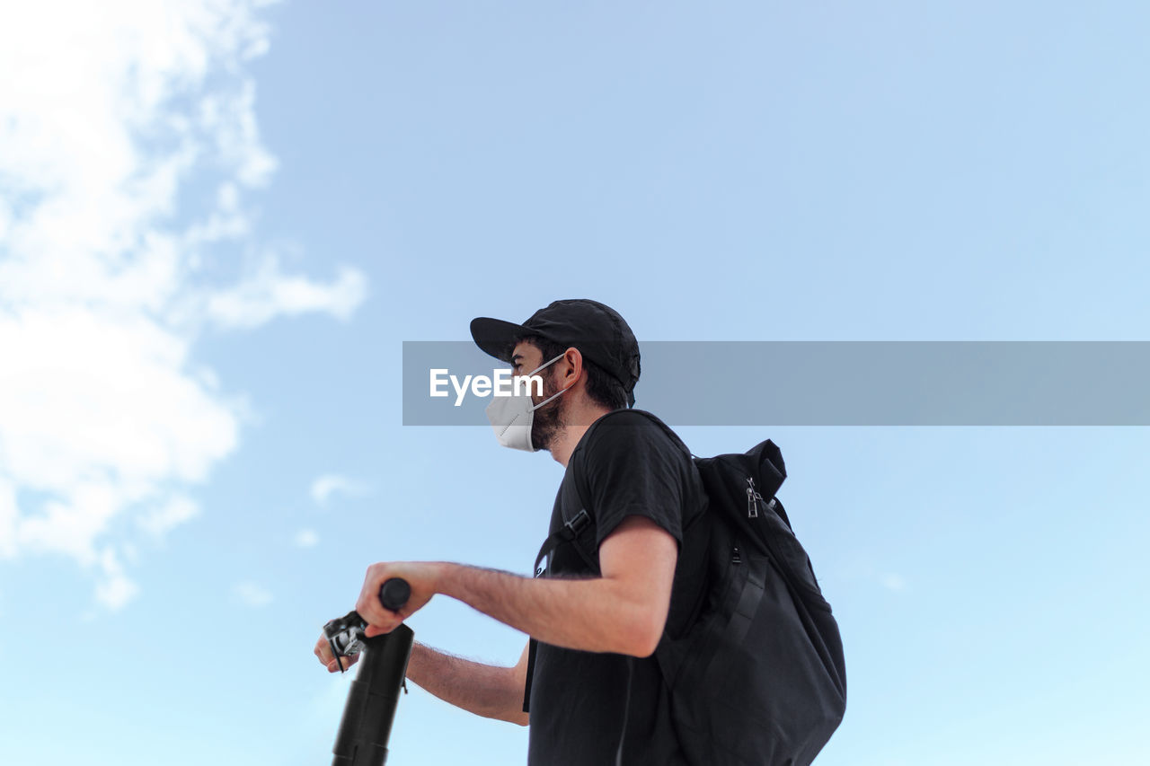 Low angle side view of anonymous male in respiratory mask and black clothes driving electric scooter under blue cloudy sky while looking forward