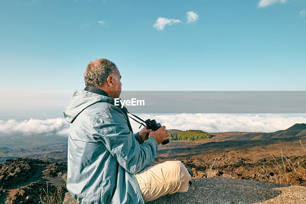 Rear view of man looking through binoculars at panoramic view of summits of active volcano etna