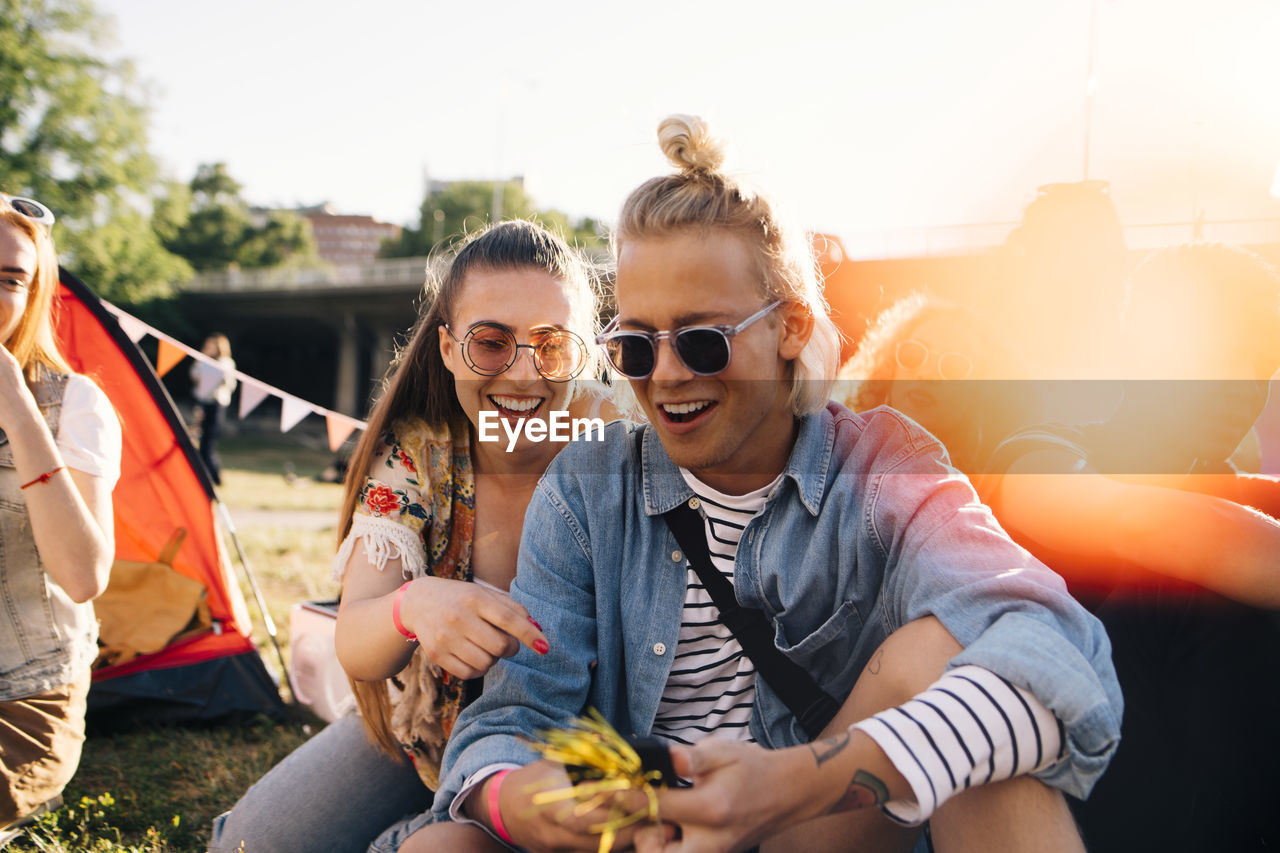 Smiling male and female friends looking at smart phone while sitting on lawn in sunny day