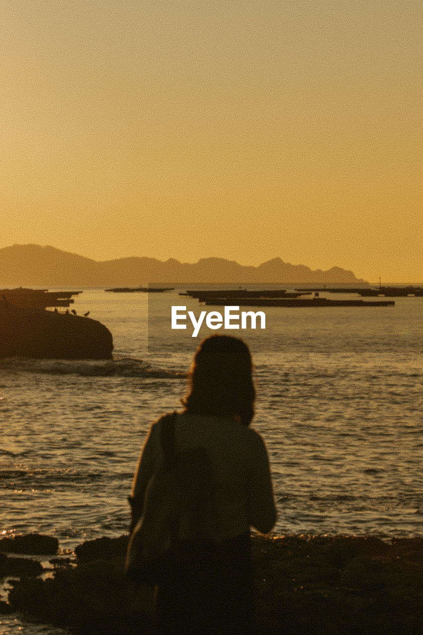 rear view of silhouette woman sitting on beach against sky during sunset