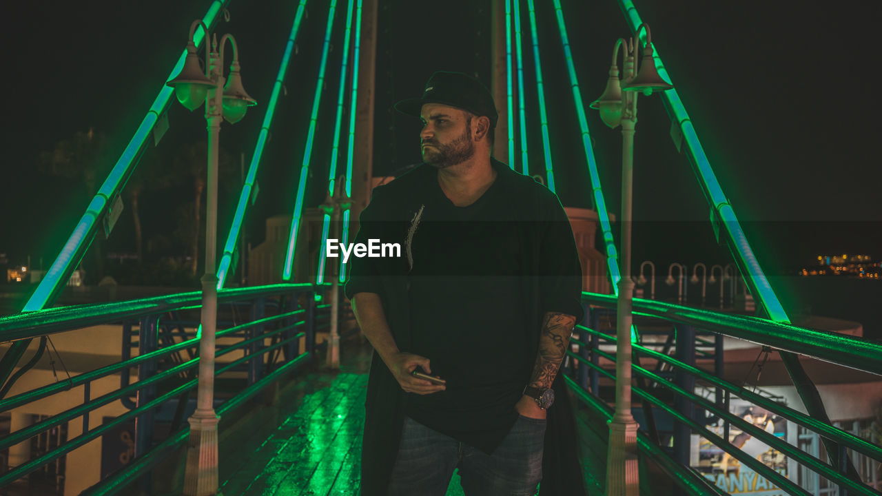 PORTRAIT OF YOUNG MAN STANDING ON ILLUMINATED BRIDGE