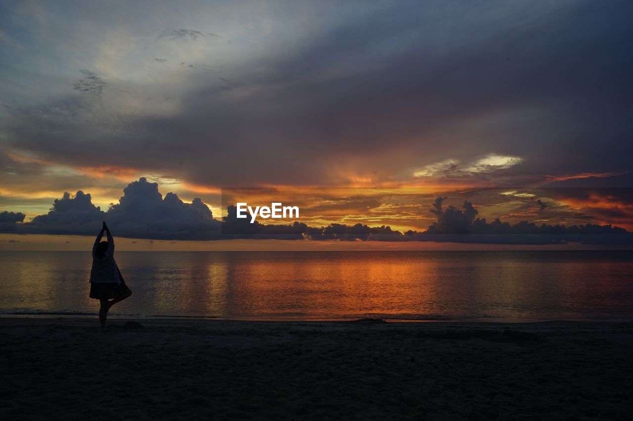 Silhouette woman doing tree pose while standing at beach against sky during sunset