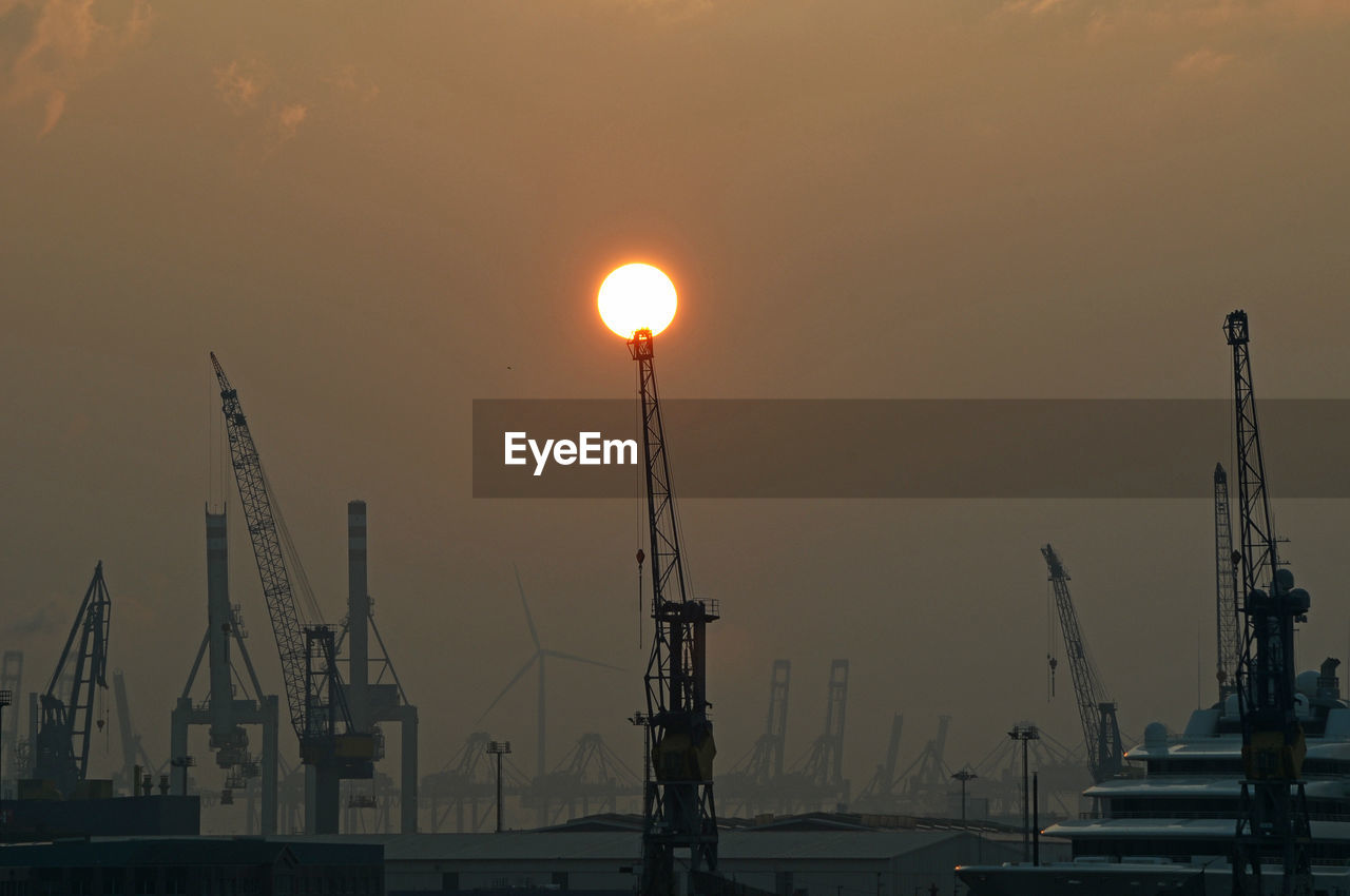 Cranes at harbor against sky during sunset