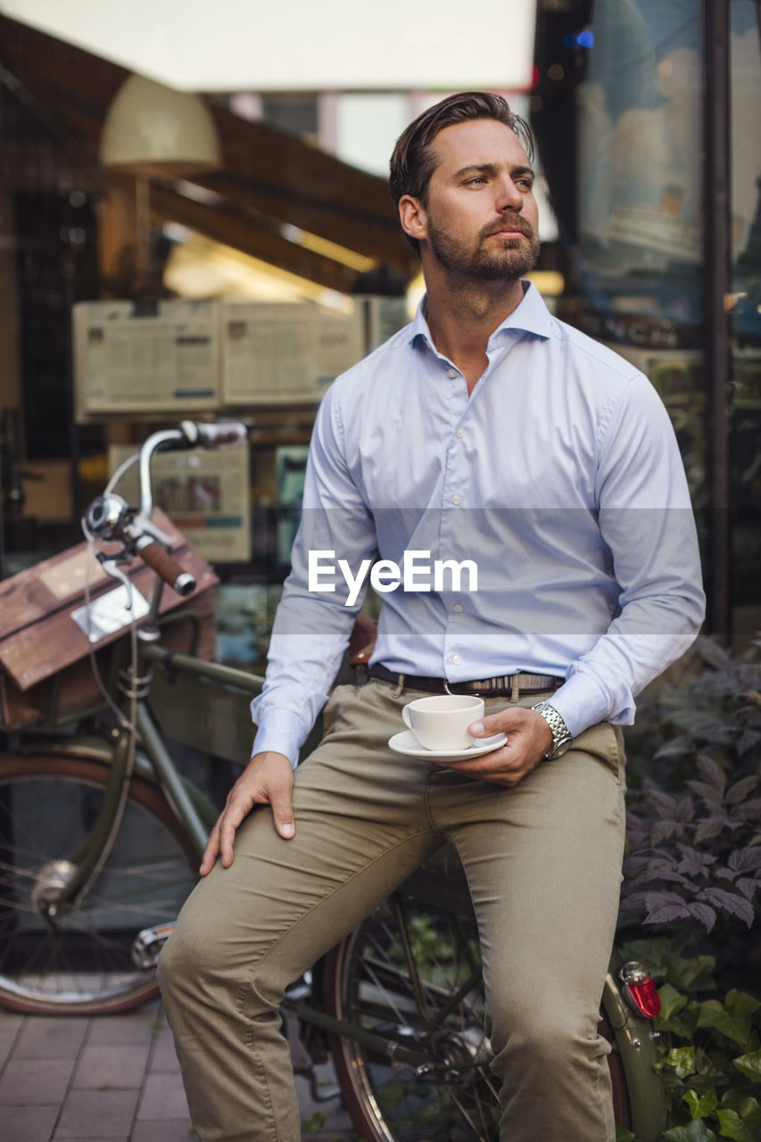 Thoughtful businessman holding coffee cup while sitting on bicycle at sidewalk cafe