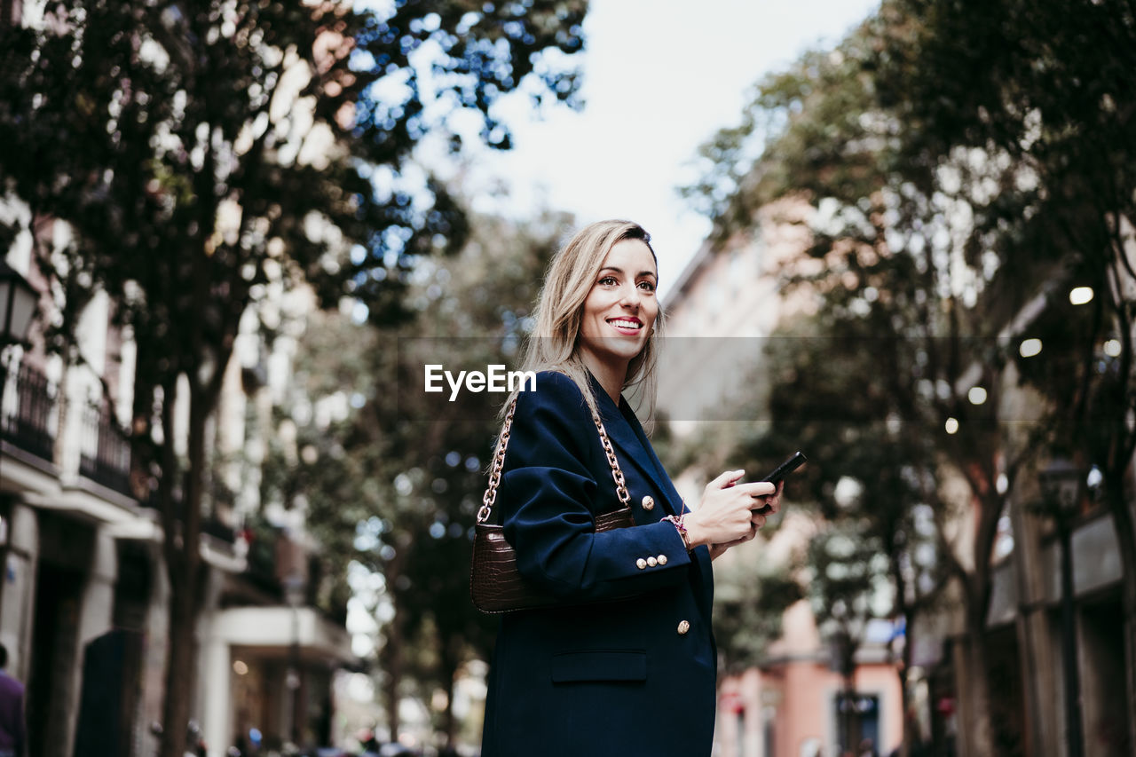 Happy beautiful woman looking away while holding mobile phone in city