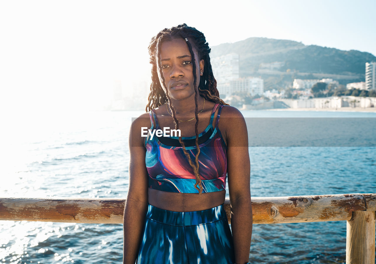 Front view of serious african american female with long dreadlocks wearing activewear standing near wooden fence on seafront during fitness workout and looking away