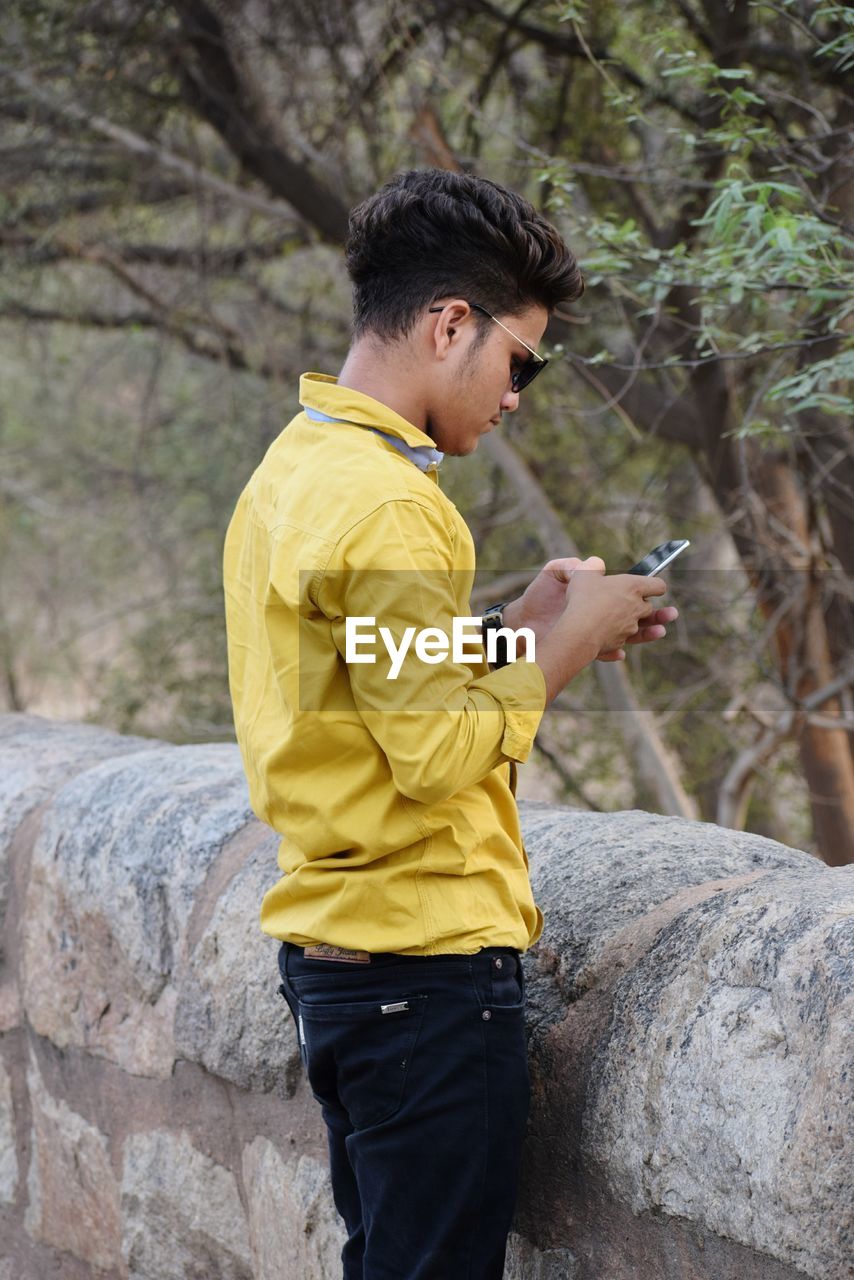 Side view of young man using phone while standing against retaining wall