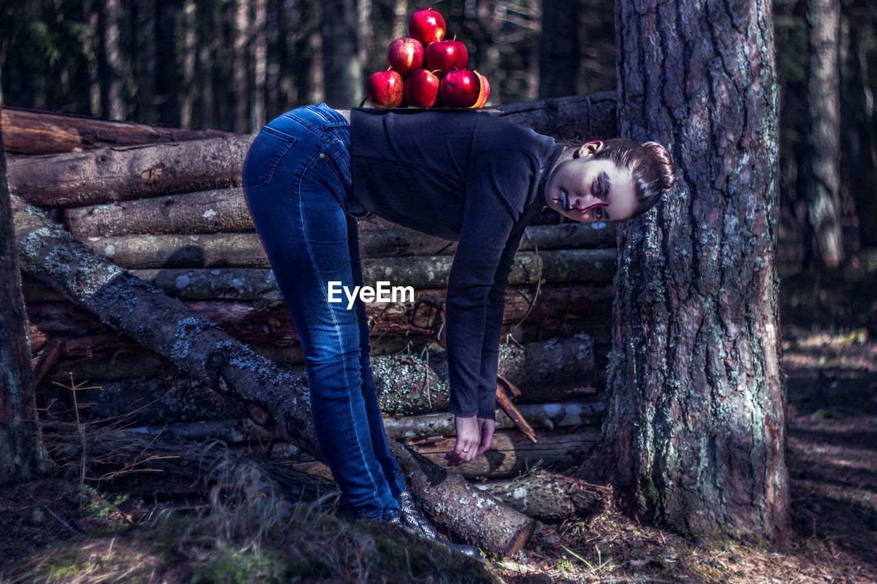Full length of woman bending with apples on back by tree trunk in forest
