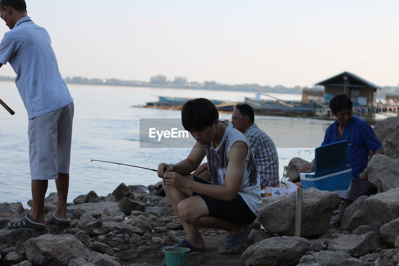 PEOPLE SITTING ON ROCKS BY SEA AGAINST CLEAR SKY