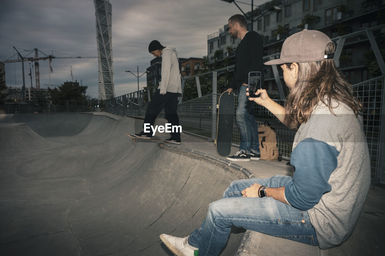 Male skateboarder skating while friend photographing on smart phone at park