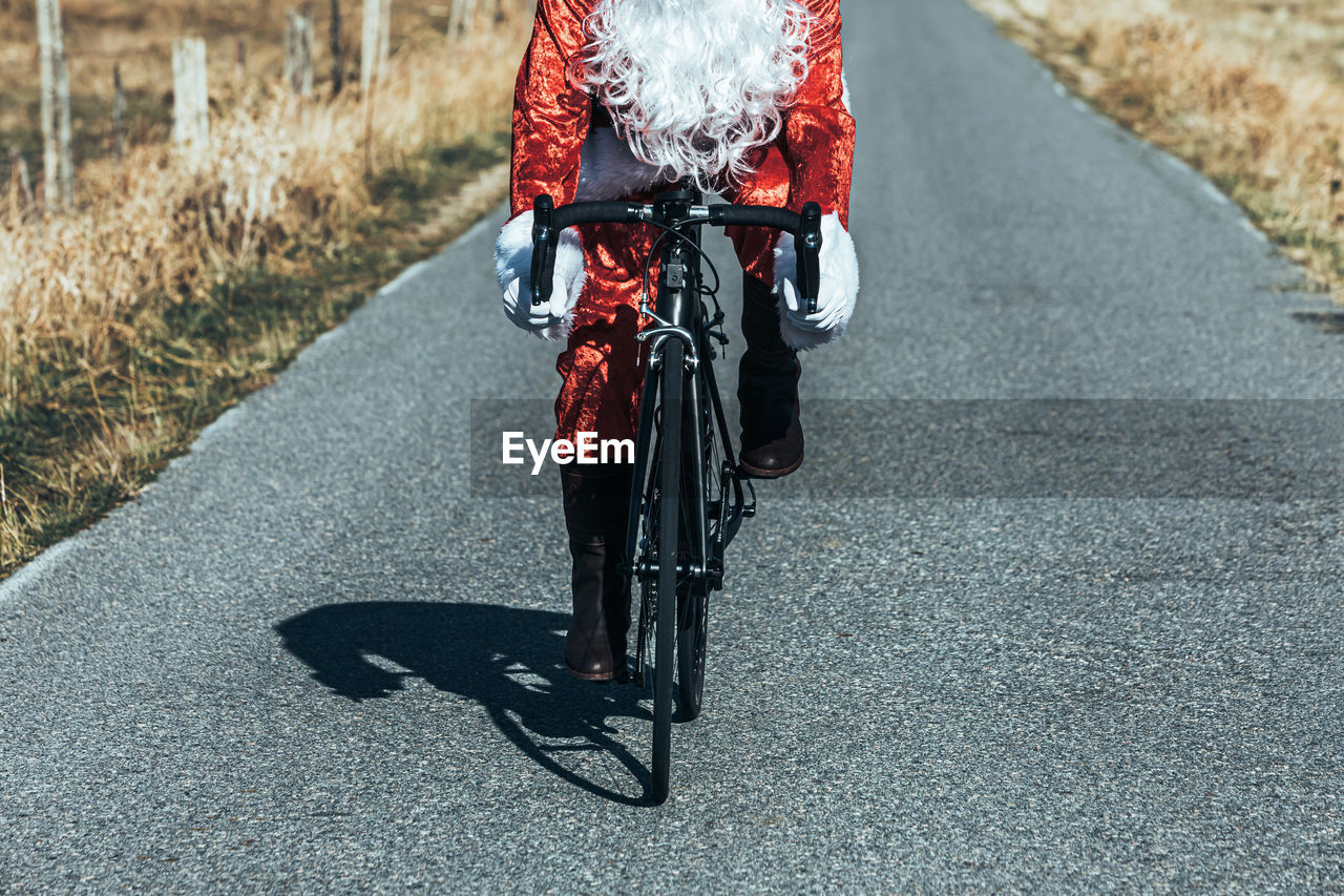Cropped unrecognizable santa claus in red costume riding modern bike along empty road in highlands on sunny day