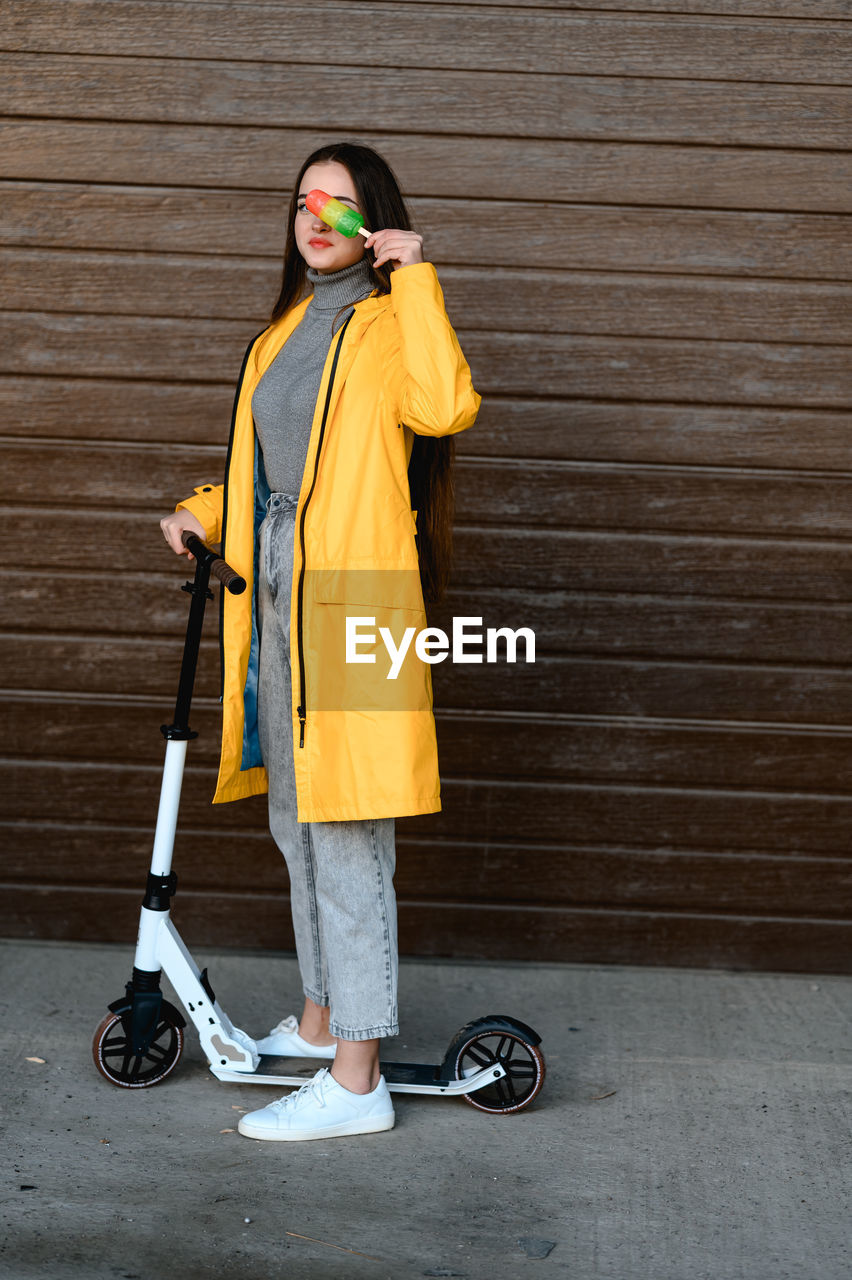 A girl in a yellow raincoat stands on a white scooter and holds colorful ice cream in her hands