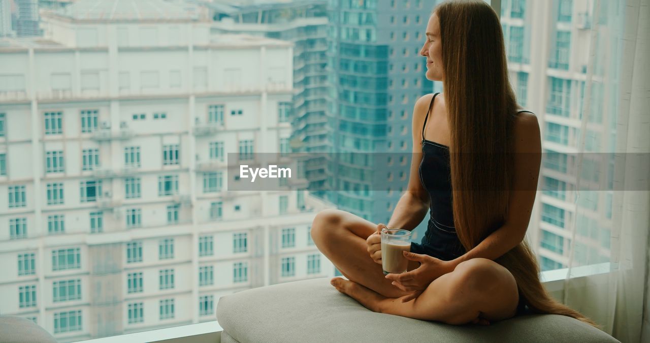 Woman drinking delicious coffee near window with big city skyscrapers outside.