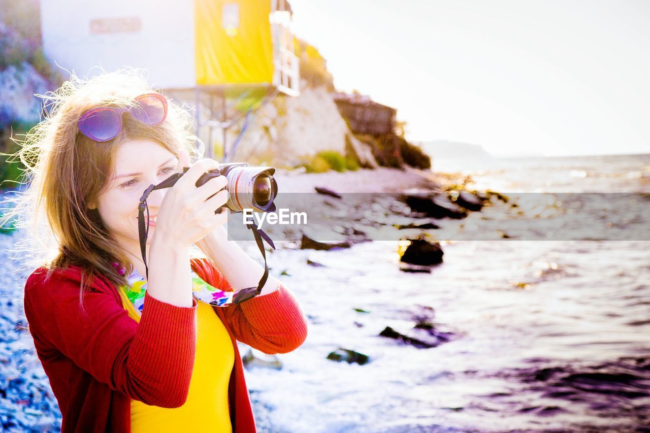 Smiling young woman photographing through digital camera by sea