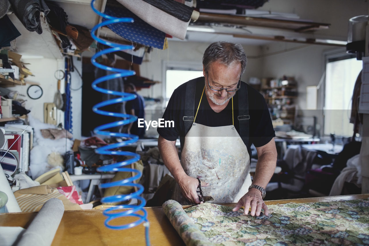 Senior male owner cutting fabric at workbench in workshop