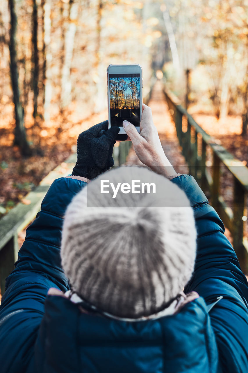 Back view of young woman taking photos of forest landscape with smartphone during trip