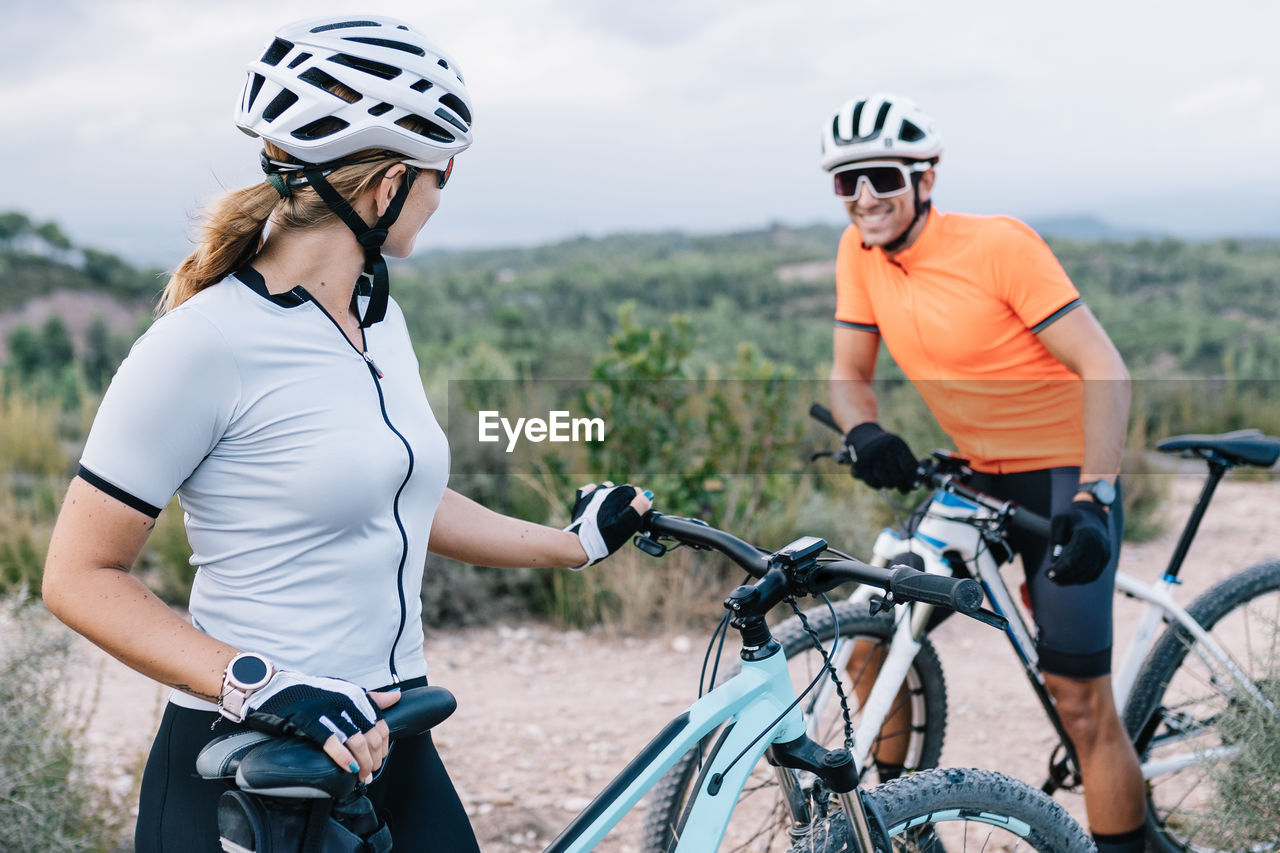 Cheerful couple of sportive bicyclists wearing helmets and sunglasses standing near modern extreme bikes and looking at each other