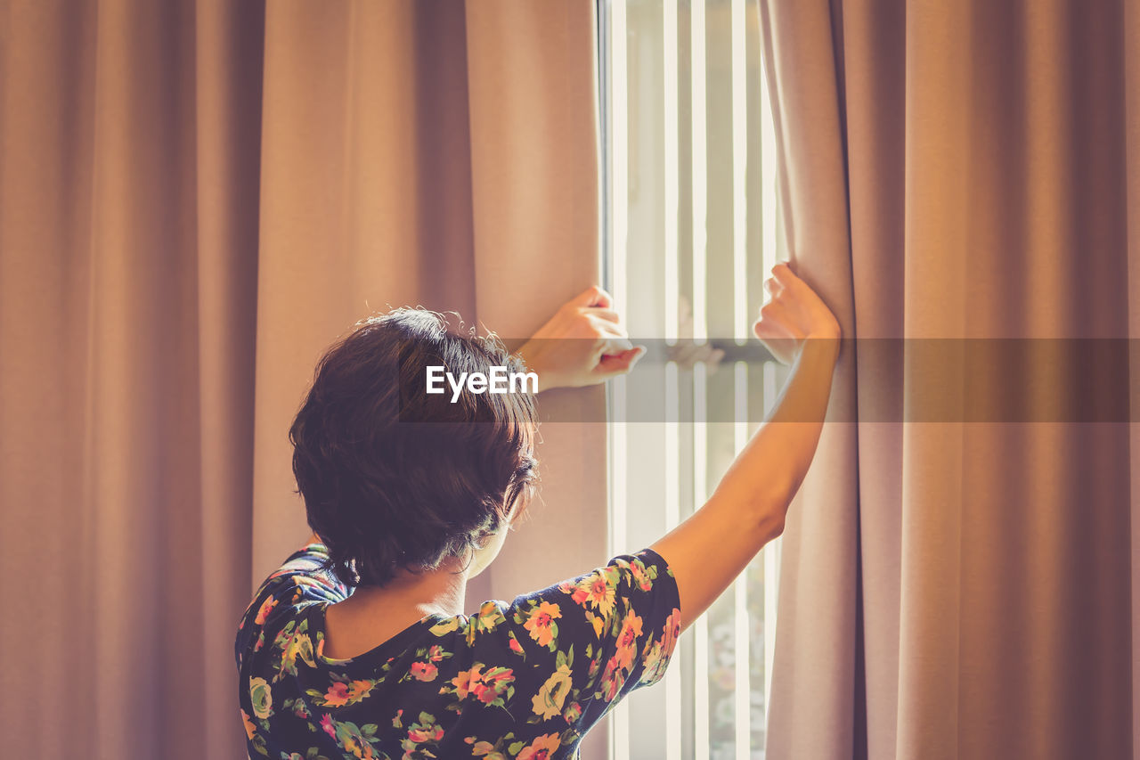 Rear view of mature woman looking through window at home