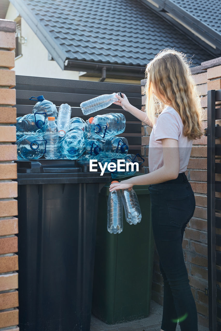 Side view of girl dumping bottles in garbage by wall