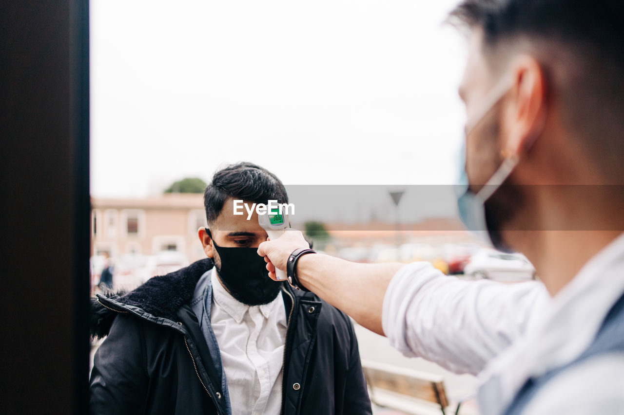 Crop anonymous male hairdresser in sterile mask measuring temperature of colleague with infrared thermometer at door of barbershop