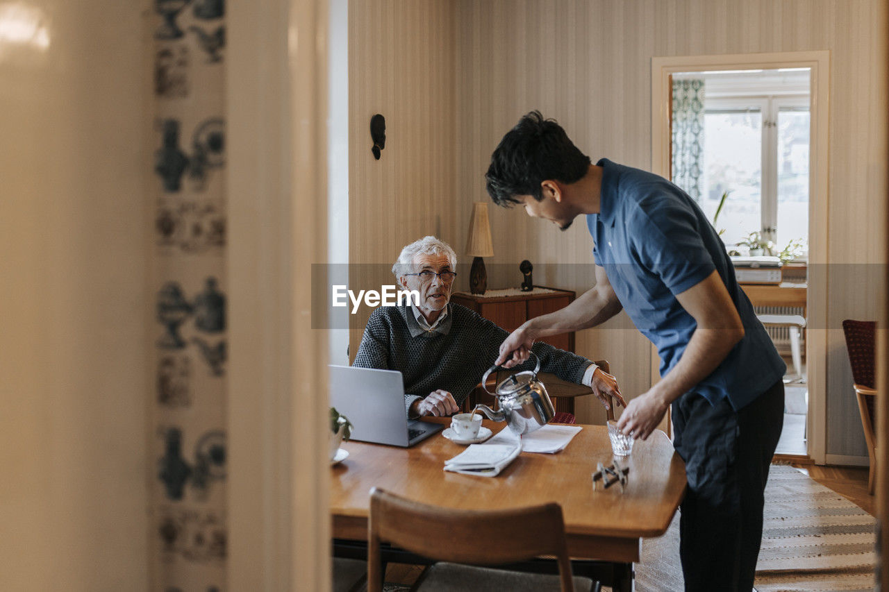 Senior man talking to male caregiver serving tea at home