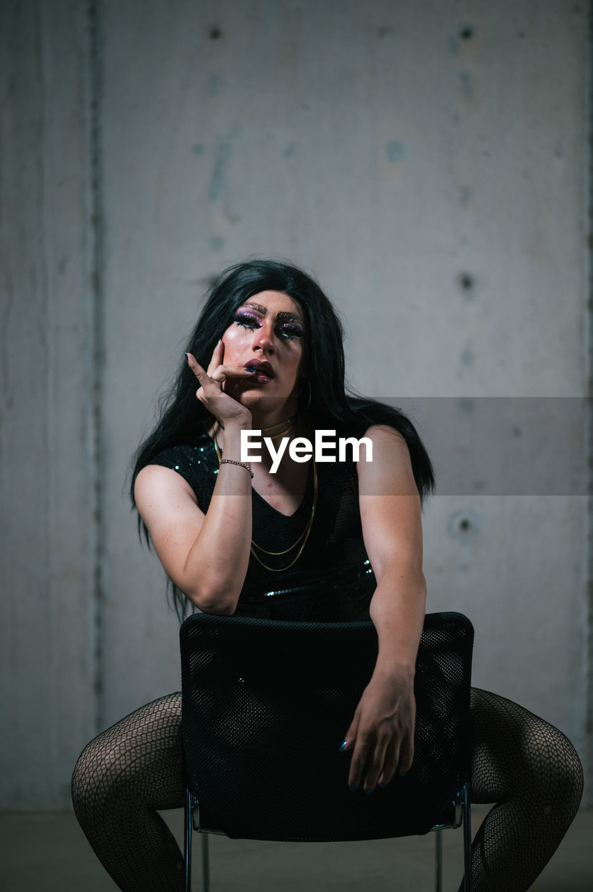 Full body of stylish drag queen wearing black garment and wig looking at camera while sitting on chair in studio